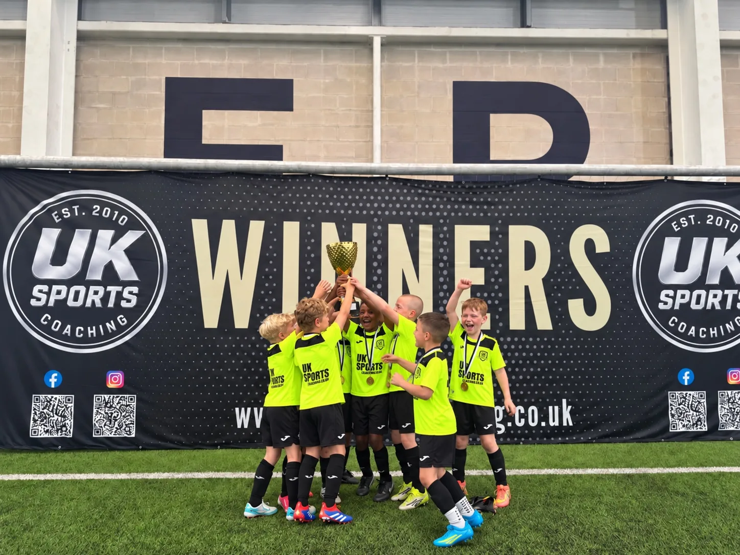 Young UK Sports team celebrating with a trophy in front of the winners backdrop